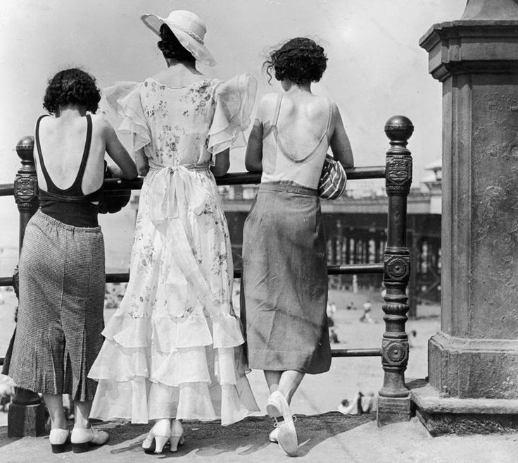 Tre donne fotografate di schiena sul lungomare, due indossano top scollati sulla schiena e gonne a tubo mentre quella al centro è vestita con un romantico abito con volant sulle maniche e in fondo alla gonna