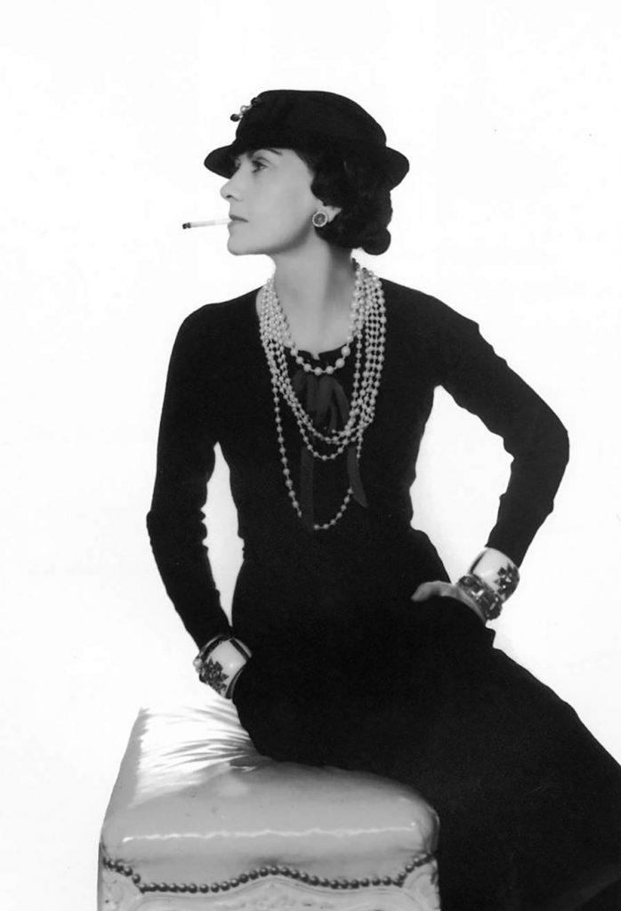 Coco Chanel fuma una sigaretta indossa un abito nero, cappello nero, collana di perle a più fili e bracciali