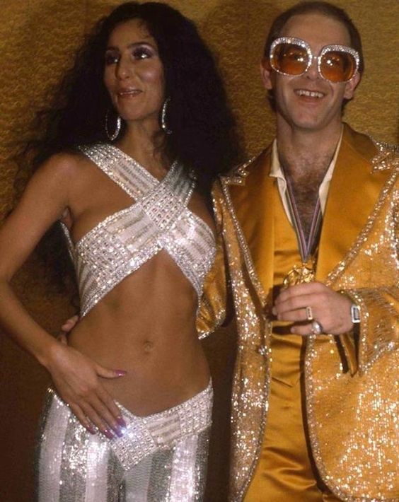 Cher indossa top e pantaloni ricoperti da paillettes bianche abbracciata a Elton John con un completo giallo oro e paillettes, 1975